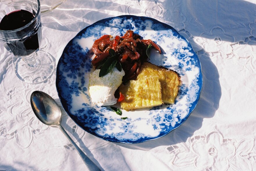 Looking down on a table with an embroidered tablecloth at a blue and white classic plate with various condiments, a spoon and a glass of red wine. 