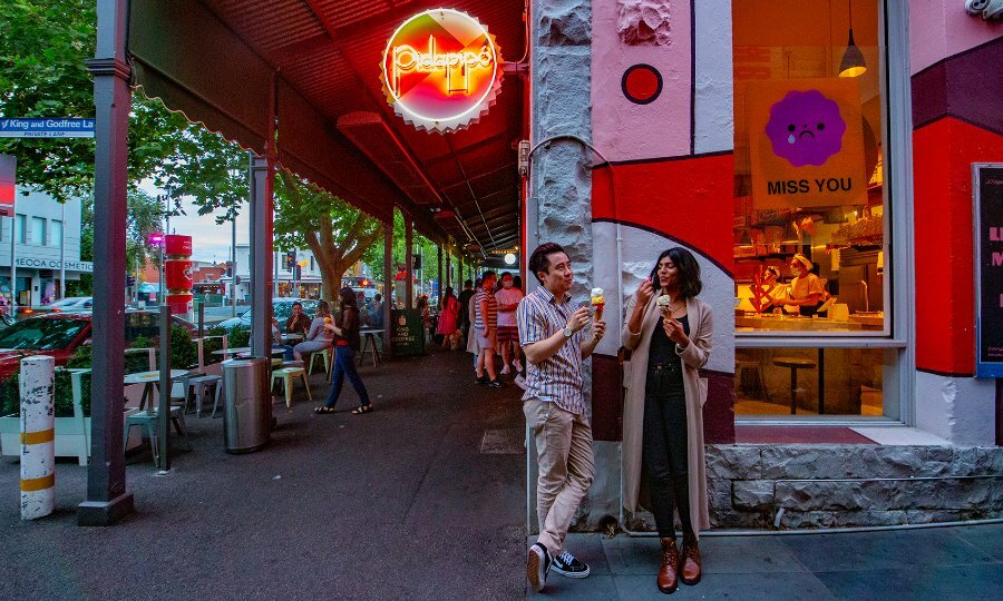 A couple leaning against the side of a colourful shop eating ice cream.