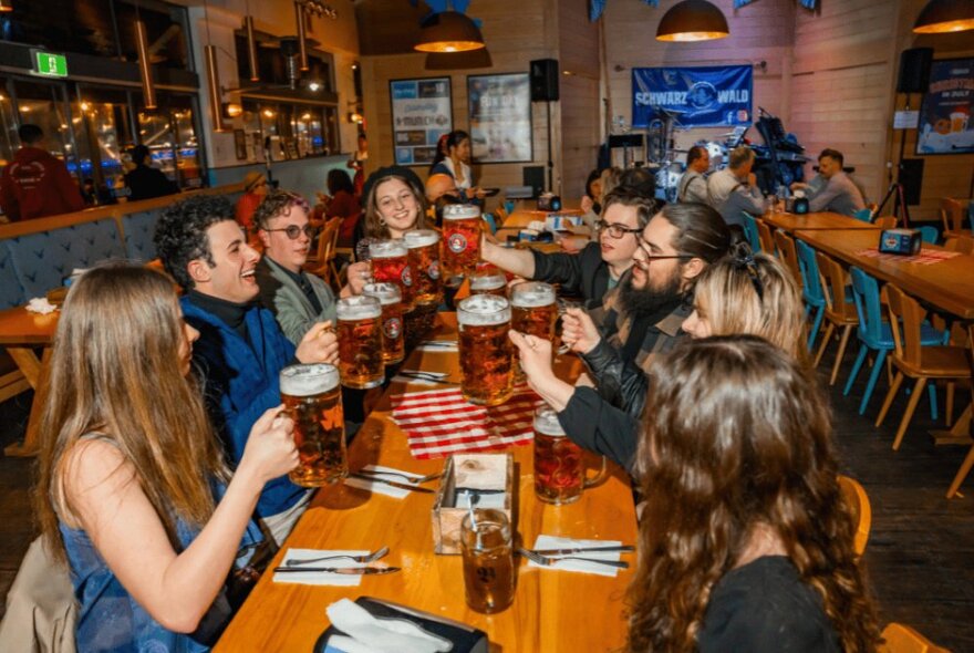 A long rectangular table in a dining hall, with people seated around the table, all holding up large steins of beer.