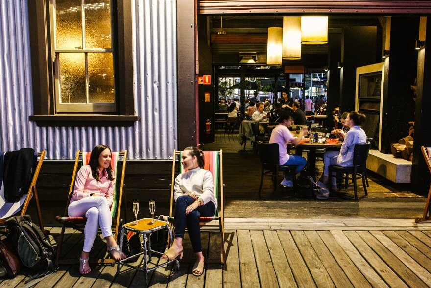 A busy restaurant with two women sitting outside on deck chairs drinking Prosecco.  