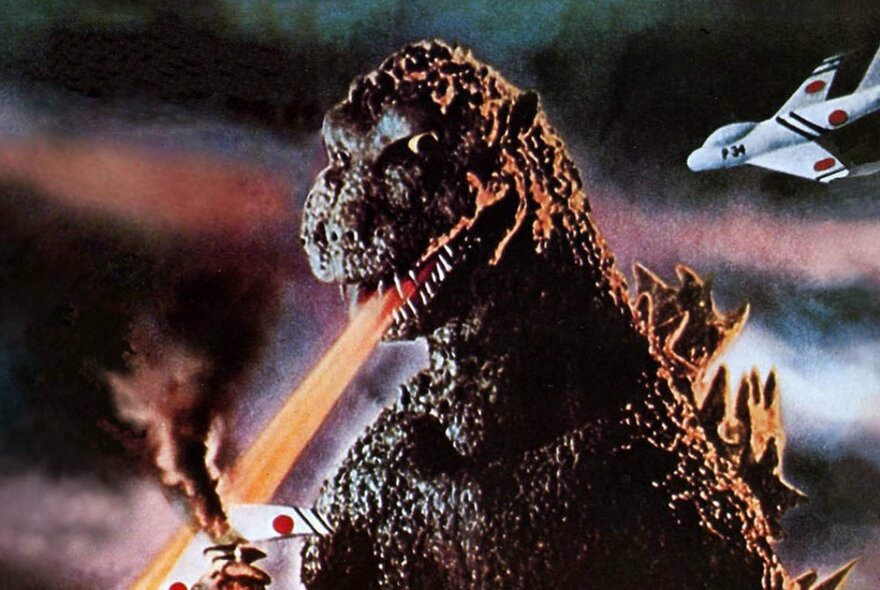 A still image of Godzilla with an orange beam coming from his mouth and a plane buzzing around behind him. 