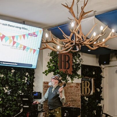Host dressed in leiderhosen and Tyrolean hat standing beneath an antler chandelier in front of a large screen.