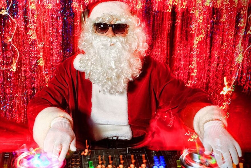 DJ dressed as Santa Claus wearing sunglasses, at music desk surrounded by red tinsel.
