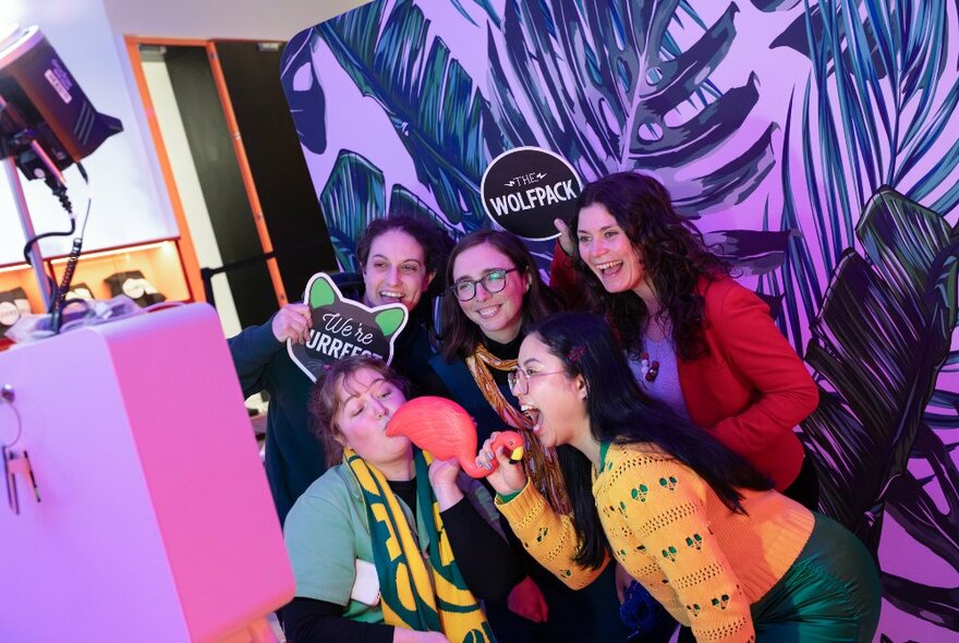 A group of young women posing for a silly photo with props in front of a purple and green background.