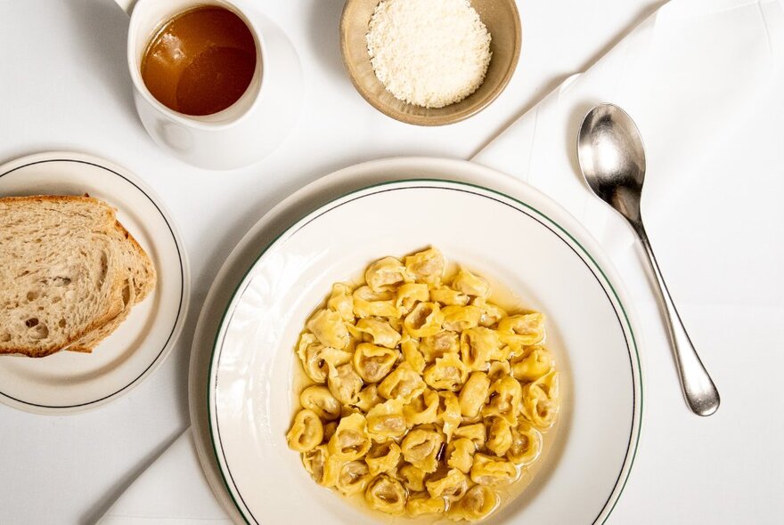 A creamy tortellini dish presented in a white bowl, a spoon, crusty bread and salt also on the table.
