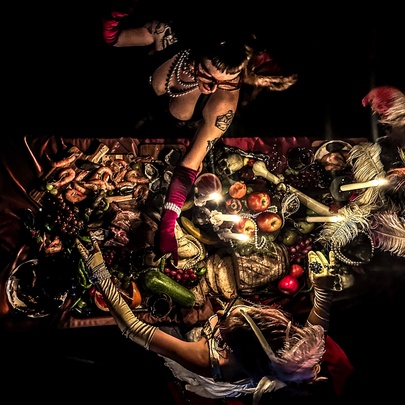 Two burlesque-clad women seen from above working over a table of food, bones and feathers.