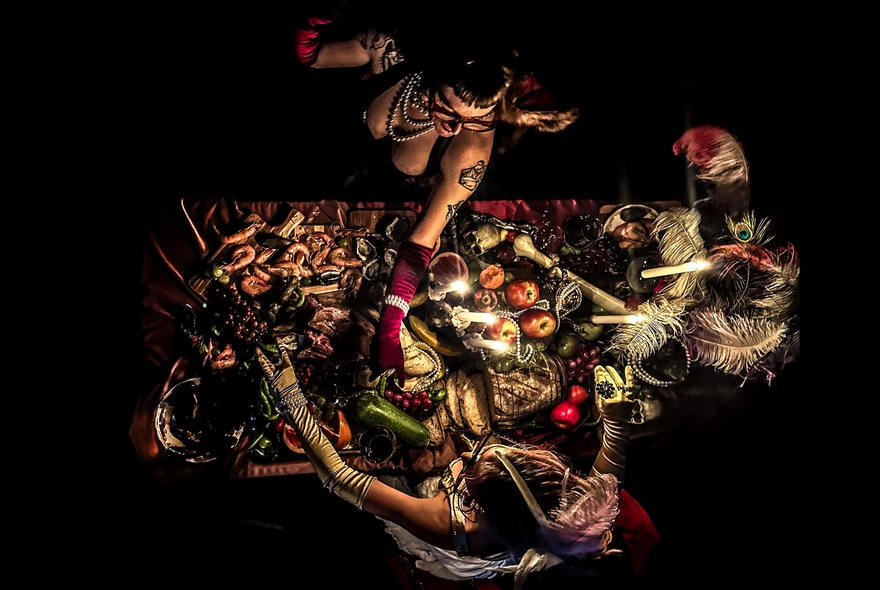 Two burlesque-clad women seen from above working over a table of food, bones and feathers.