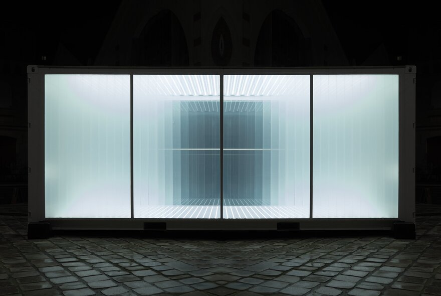 Illuminated room of shimmering white light and mirrors, set behind a black framed window, with dark bluestone pavers in the foreground.