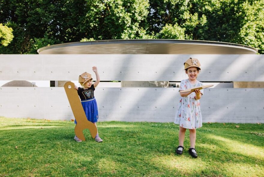 Two young children holding pieces of cardboard, standing on grass outside a concrete building.