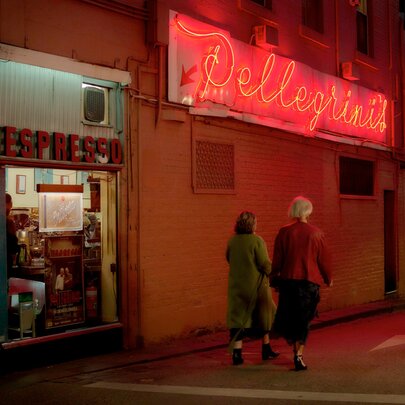 Two people walking past Pellegrini's Cafe on a winter's night, a view through the glass windows into the brightly lit cafe, the red neon cursive script Pellegrini's sign is illuminated.