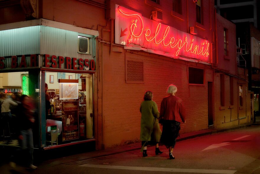 Two people walking past Pellegrini's Cafe on a winter's night, a view through the glass windows into the brightly lit cafe, the red neon cursive script Pellegrini's sign is illuminated.