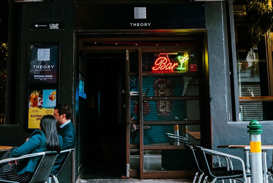 Exterior street view of Theory Bar, showing people drinking at tables outside and an open glass door.