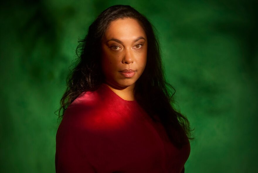 Indigenous actor with long dark hair wearing a red dress against a green background.