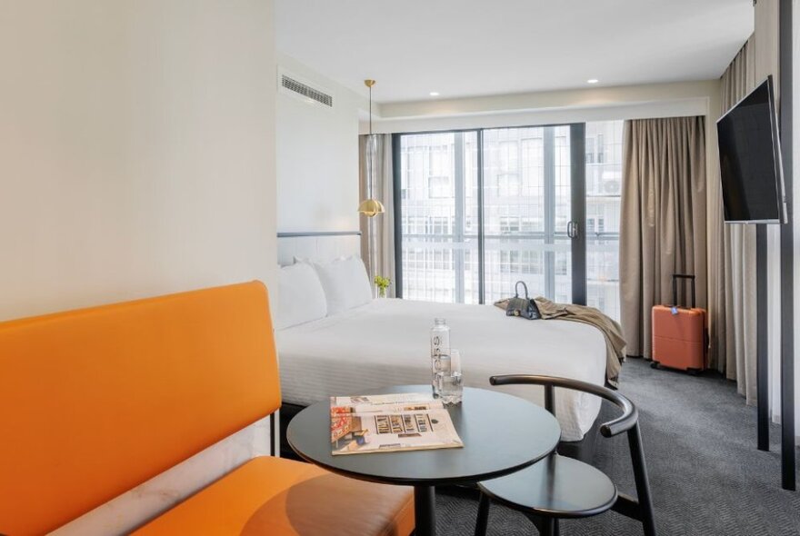 Hotel room with large double bed, orange sofa, table and chair, and large window with drapes.