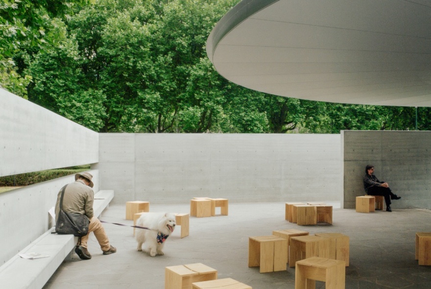 Inside MPavilion under the umbrella canopy, people sitting on small wooden stools, one with a dog on a leash. 