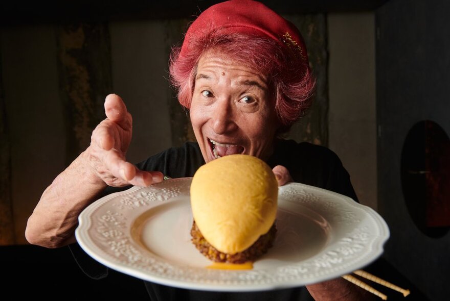 Chef with dyed red hair smiling while holding a white plate with omelet and rice, with chopsticks.