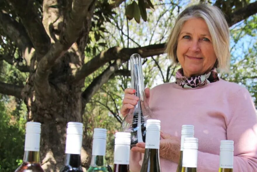 Winemaker Ros Ritchie standing outdoors and holding a carafe, the tops of several bottles of wine visible in front of her on a table.