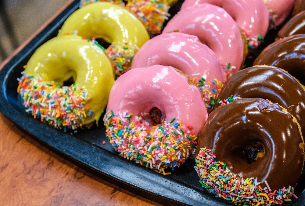 Rows of yellow, pink and brown doughnuts with sprinkles on a black plastic tray.