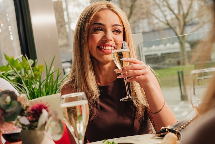 A young woman enjoying a glass of champagne in a restaurant setting; a floral arrangement on the table.