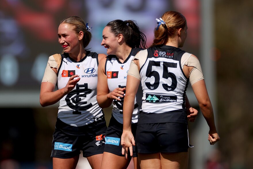 Three Carlton AFLW players celebrating and smiling on the field.
