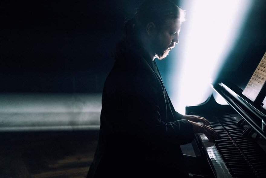 Bearded pianist in profile at the keyboard in shafts of bright light in a dark space.