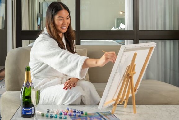 A happy woman in a white bathrobe painting on a small easel with a bottle of champagne open beside her.