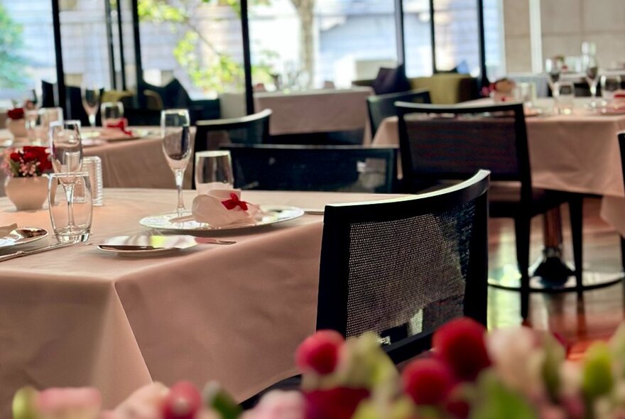 A dining room with square tables covered in white linen tablecloths, set for a meal service with crockery, cutlery and glassware. 