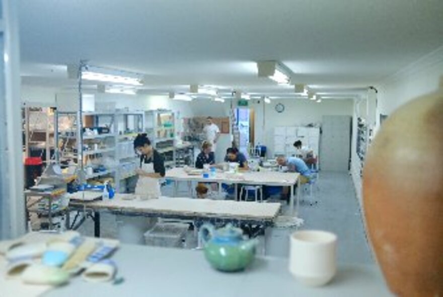 A brightly lit pottery studio with people creating at long tables and various pottery items around the room. 