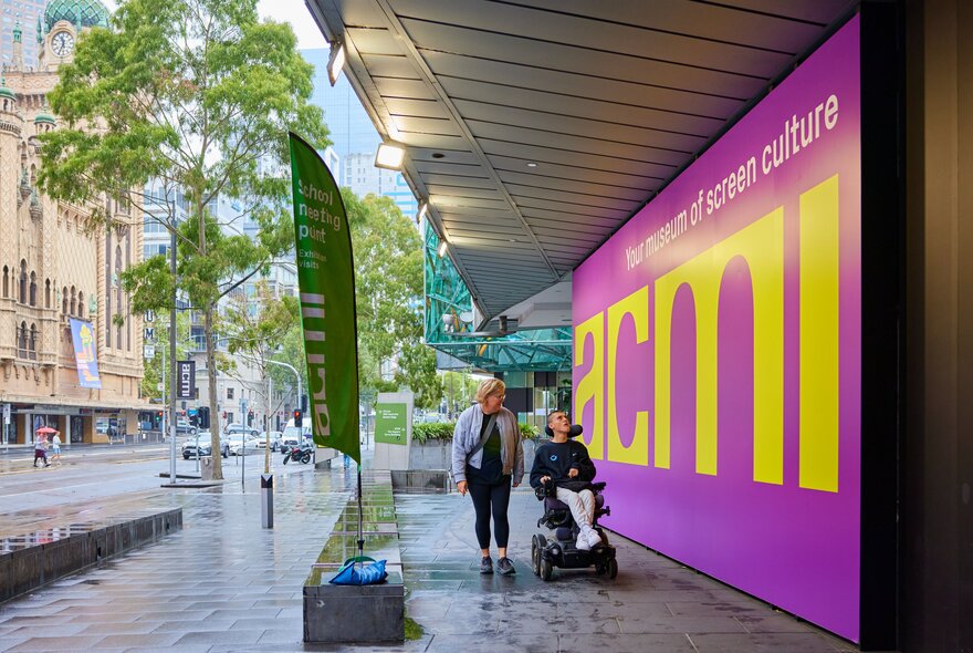 A man in a powerchair and woman strolling past a big purple and yellow sign with the word "acmi" on it.