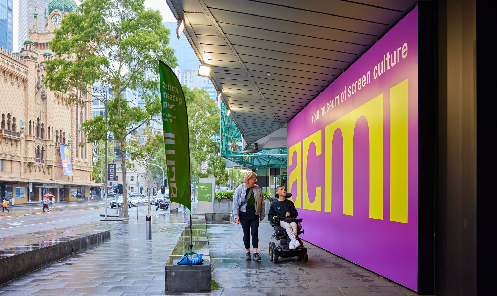 A man in a powerchair and woman strolling past a big purple and yellow sign with the word "acmi" on it.