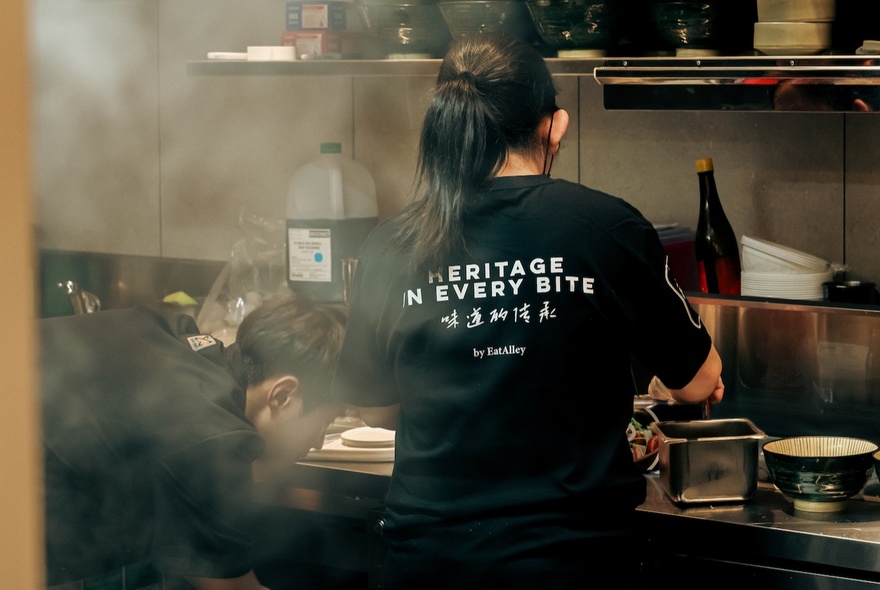The back of a person wearing a black t-shirt cooking in a commercial kitchen, with kitchen pans and implements around them and steam in the foreground. 