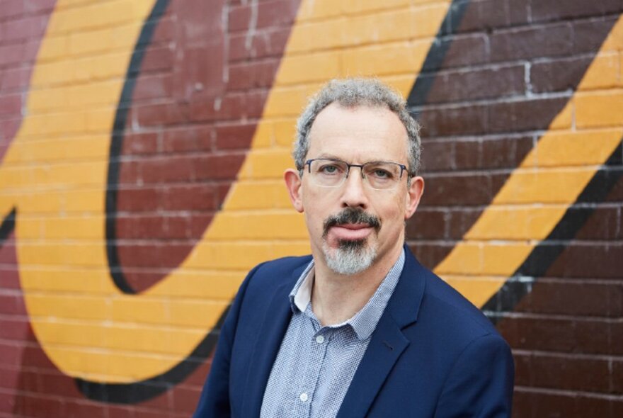 Upper half of the body of a man wearing a navy suit jacket and pale shirt, with a grey beard and short hair, glasses on his face, posed against a painted brick wall.