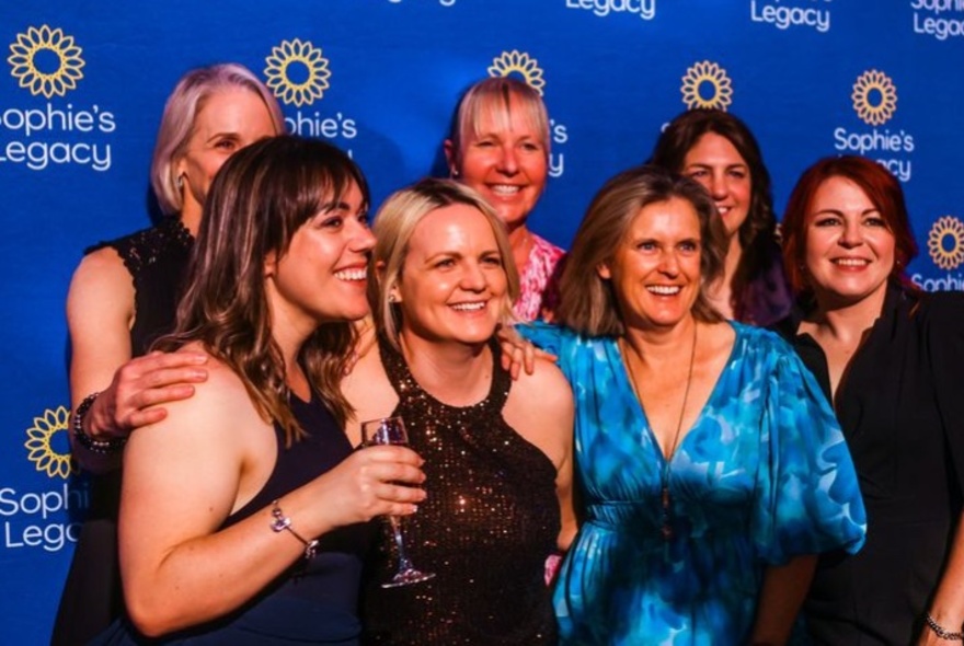 A group of smiling people standing in front of a branded blue backdrop for Sophie's Legacy, an organization that advocates for the wellbeing and mental health of veterinary professionals.