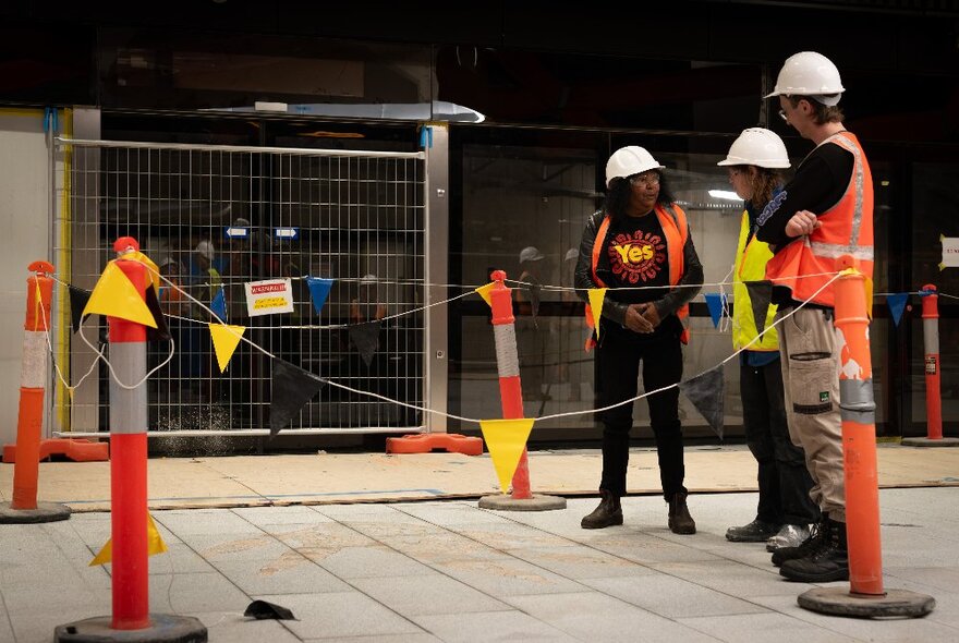 Three people wearing high vis vests and hard hats standing at a construction site and talking to each other.