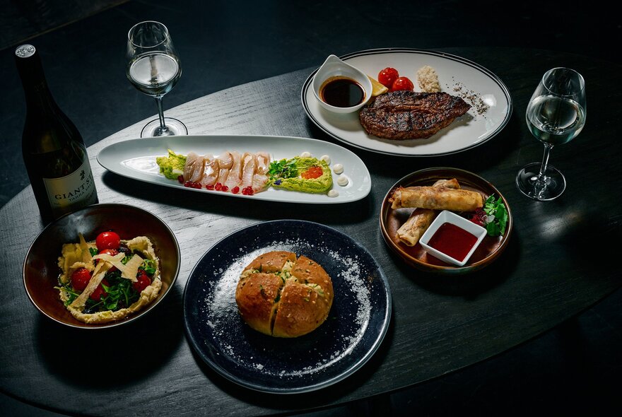 A dark-toned table in a restaurant with several plates of food, including grilled steak, sliced fish tartare, a salad, baked bread, spring rolls, two glasses of white wine, and a bottle of wine.