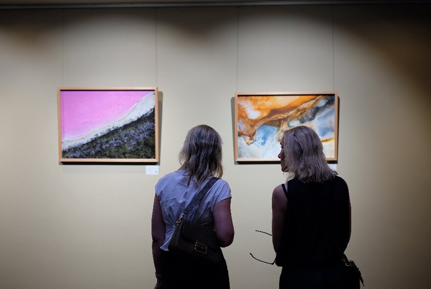 Two people looking at artworks hanging on a dimly lit grey wall in a gallery.