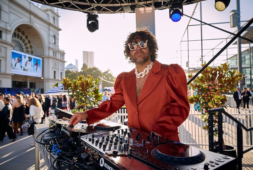 A DJ behind a mixing console at Museum Plaza with a crowd of people gathered in front of the Royal Exhibition Building; daytime.