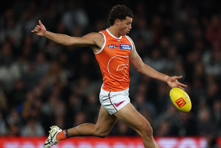 A GWS Giants AFL football player running and just about to kick the footy, a blurred stadium crowd in the background.