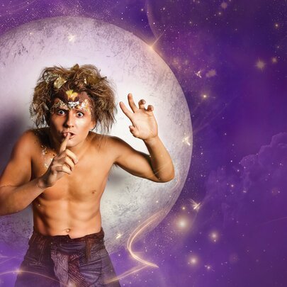 A performer dressed as Puck from A Midsummer Night's Dream, shirtless and standing in front of a purple celestial sky with a large moon as he makes the 'shh' gesture.