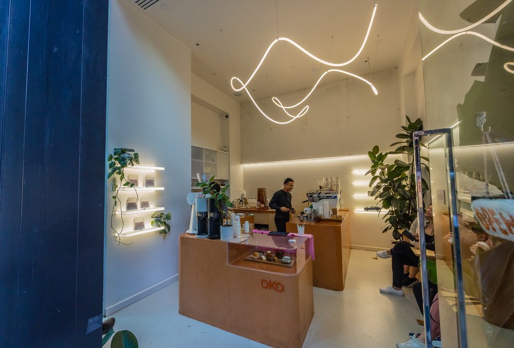 A barista in a small cafe with neon lights and plants.