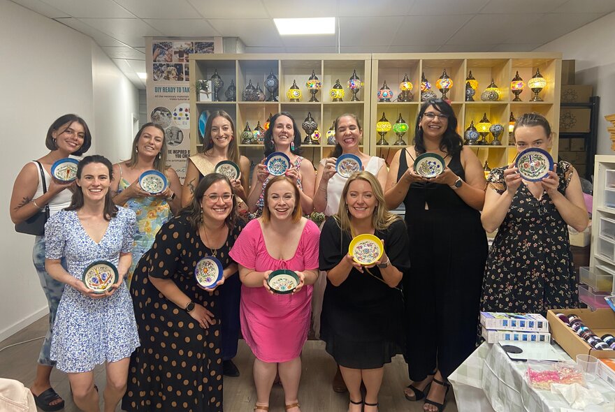 A large group of women smiling and holding up painted bowls at a craft workshop. 