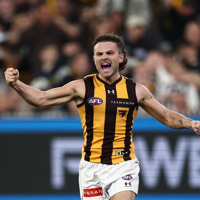A Hawthorn AFL football player with clenched fists running on the football field and cheering, a blurred stadium crowd of fans in the background.