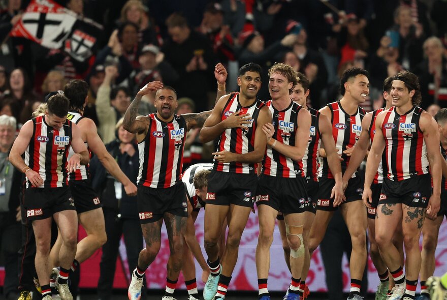 Players from the St Kilda football team looking happy after a win on the football field with a crowd in the background. 