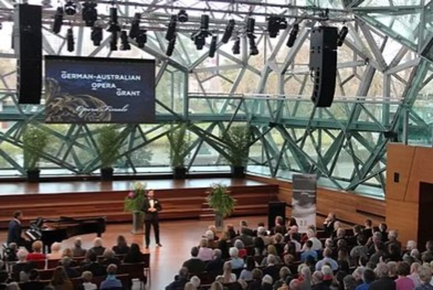 Event in the Edge with tuxedo-wearing presenter on stage in front of seated audience.
