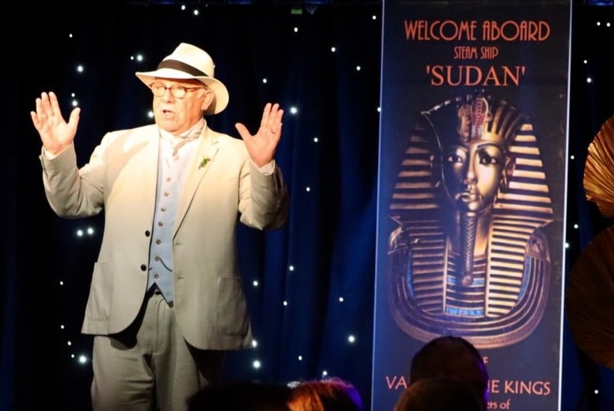 An actor dressed in a light coloured linen suit, hosting a whodunnit dinner, talking in front of a poster that says 'Welcome aboard Sudan'.