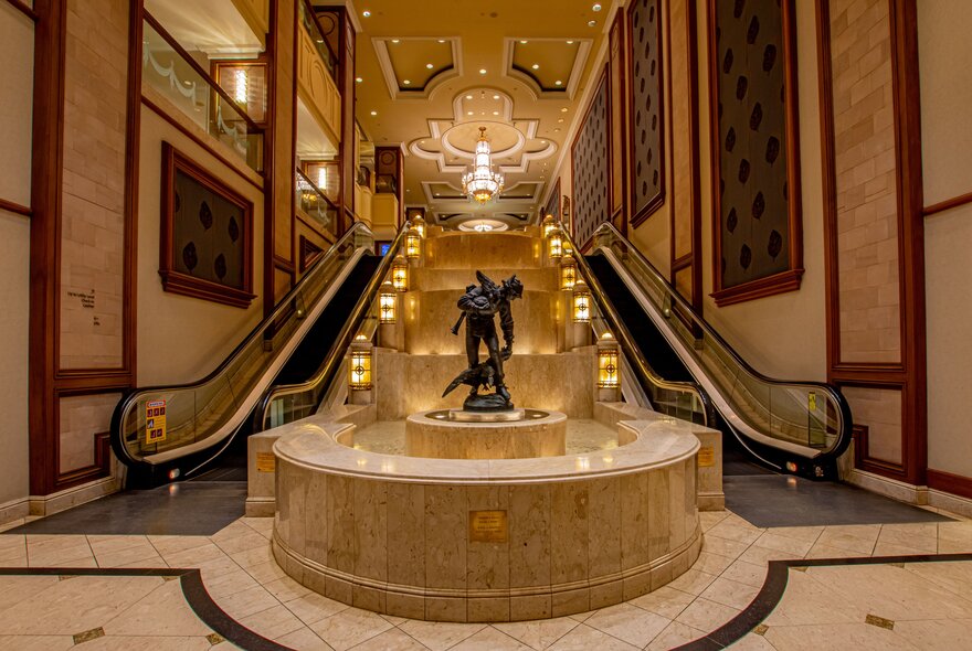 The grand entrance lobby of The Langham, Melbourne, featuring a cascading water feature and escalators leading up to the main reception area. 