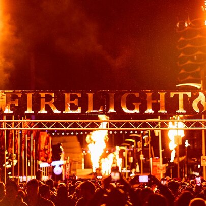 Large lettering saying FIRELIGHT above a stage at the Firelight Festival with two large flames shooting into the air at either end. 