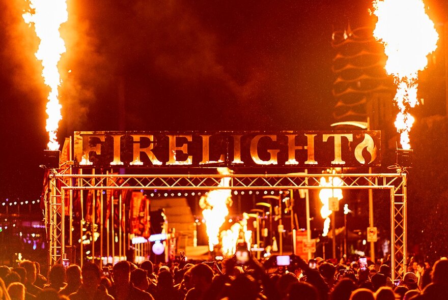 Large lettering saying FIRELIGHT above a stage at the Firelight Festival with two large flames shooting into the air at either end. 