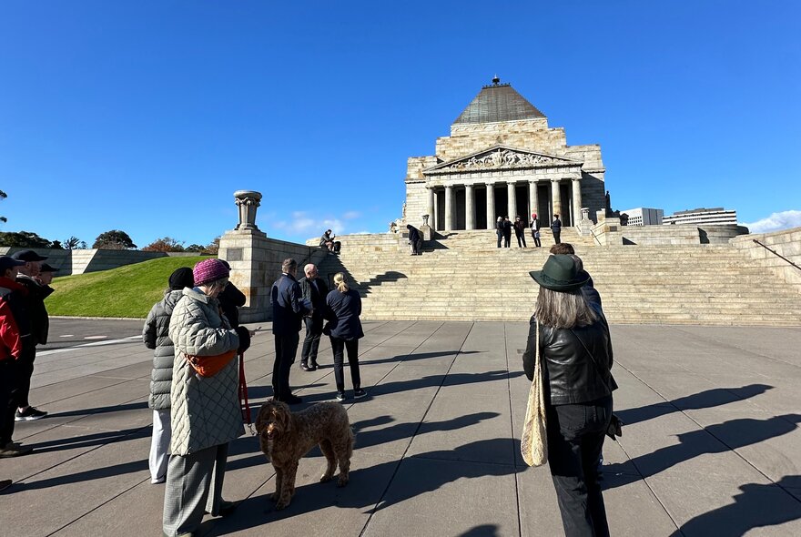 A group of people and a dog stand in the paved foreground of the Shrine of Remembrance on a sunny day listening to a tour leader.