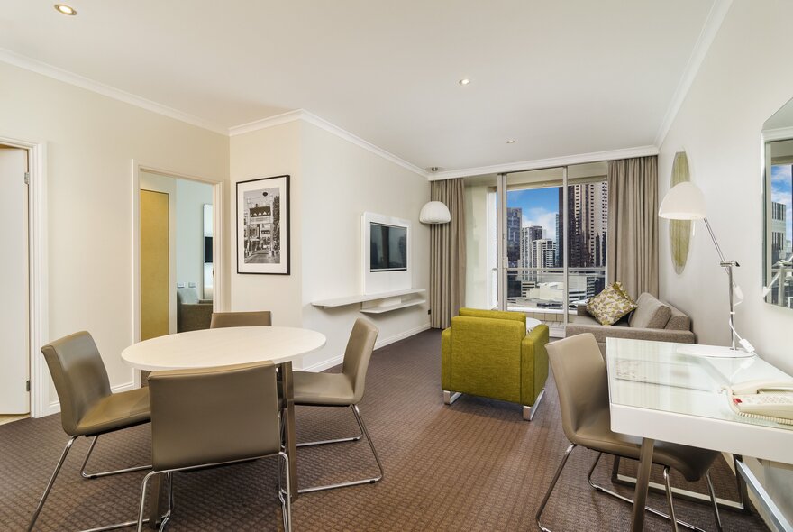 Hotel suite with lounge and dining furniture in front of window looking onto city views.